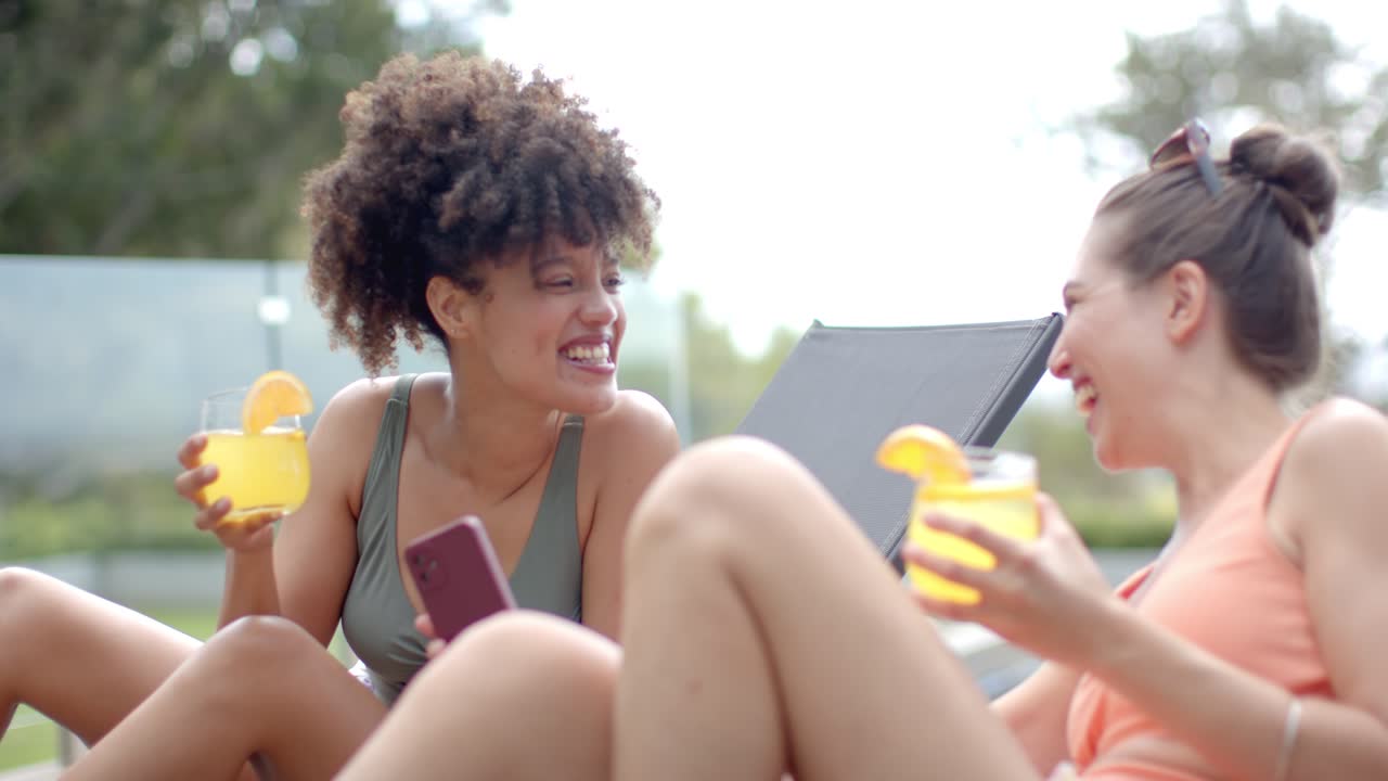 Diverse amiche che reagiscono al telefono, con bevande arancioni, occhiali da sole in testa, che ridono a bordo piscina.