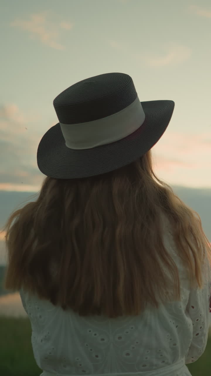 색 드레스를 입고 머리카락과 검은 모자에 초점을 맞추는 여성의 가까운 뒤면, 그녀는 조용한 풀에서 혼자 서서, 팔을 고, 평화로운 일몰 동안 조용한 호수를 쳐다보고 있습니다.