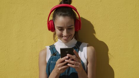 Joven-Y-Atractiva-Mujer-Latina-Que-Escucha-Música-Con-Auriculares-Inalámbricos-Recibe-Un-Mensaje-Móvil-Y-Sonríe-De-Pie-Contra-Una-Pared-Amarilla-En-Un-Día-Soleado-Tiro-Medio