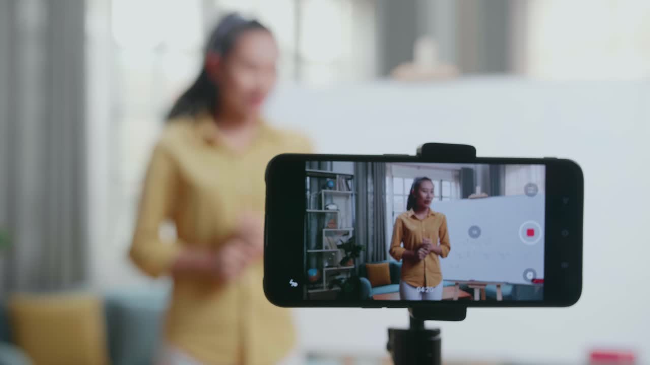 Nærbillede af smartphone-kamera, der optager asiatisk kvindelig lærer med en tuschpen, der underviser i matematik hjemme.