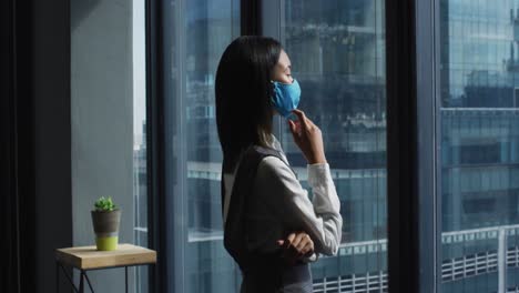 mujer asiática pensativa con máscara facial mirando por la ventana en una oficina moderna