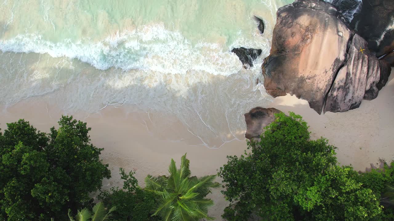 Bird eye drone shot of white sandy beach, large granite stone, coconut palm trees and waves crashing on the shore, Mahe Seychelles 60 fps 1