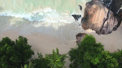 bird eye drone shot of white sandy beach, large granite stone, coconut palm trees and waves crashing on the shore, mahe seychelles 60 fps 1