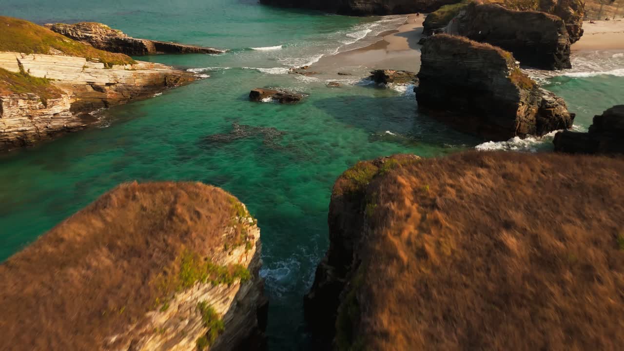 Aerial flyover of rugged coastal cliffs with natural stone arches and turquoise waters below, leading towards sandy beaches and distant hills