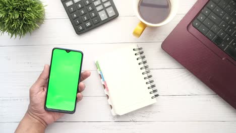 hand holding smart phone with green screen with laptop on office desk
