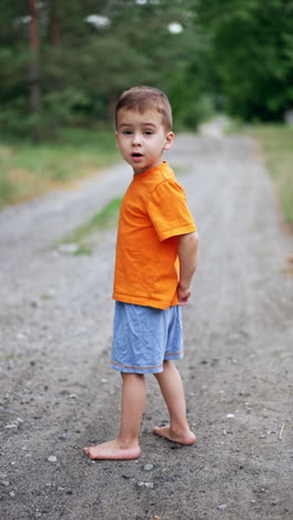 caucasian toddler in orange t-shirt and blue shorts stands barefoot on the ground. a walk in the nature in summer. vertical video.