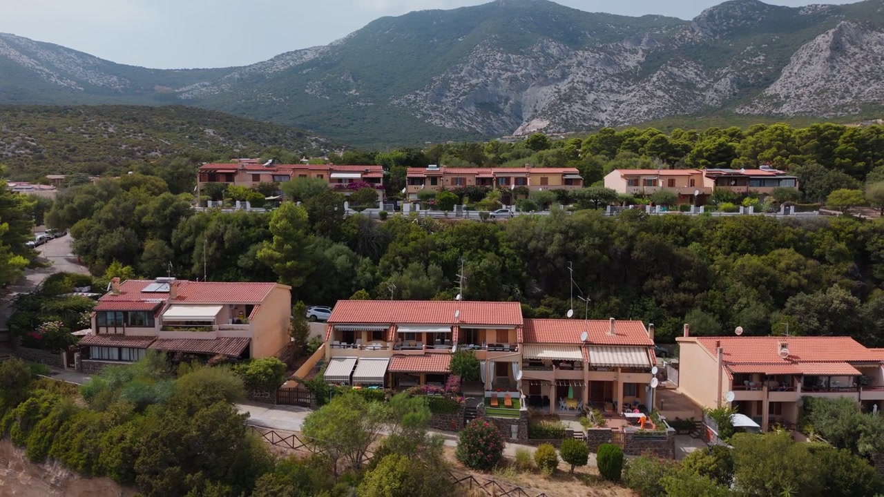 Case costiere con alle spalle aspre montagne vicino alla spiaggia di Palmasera, Cala Gonone, Sardegna, Italia. Ideale per temi di viaggio, turismo e stile di vita mediterraneo, vista aerea cinematografica.