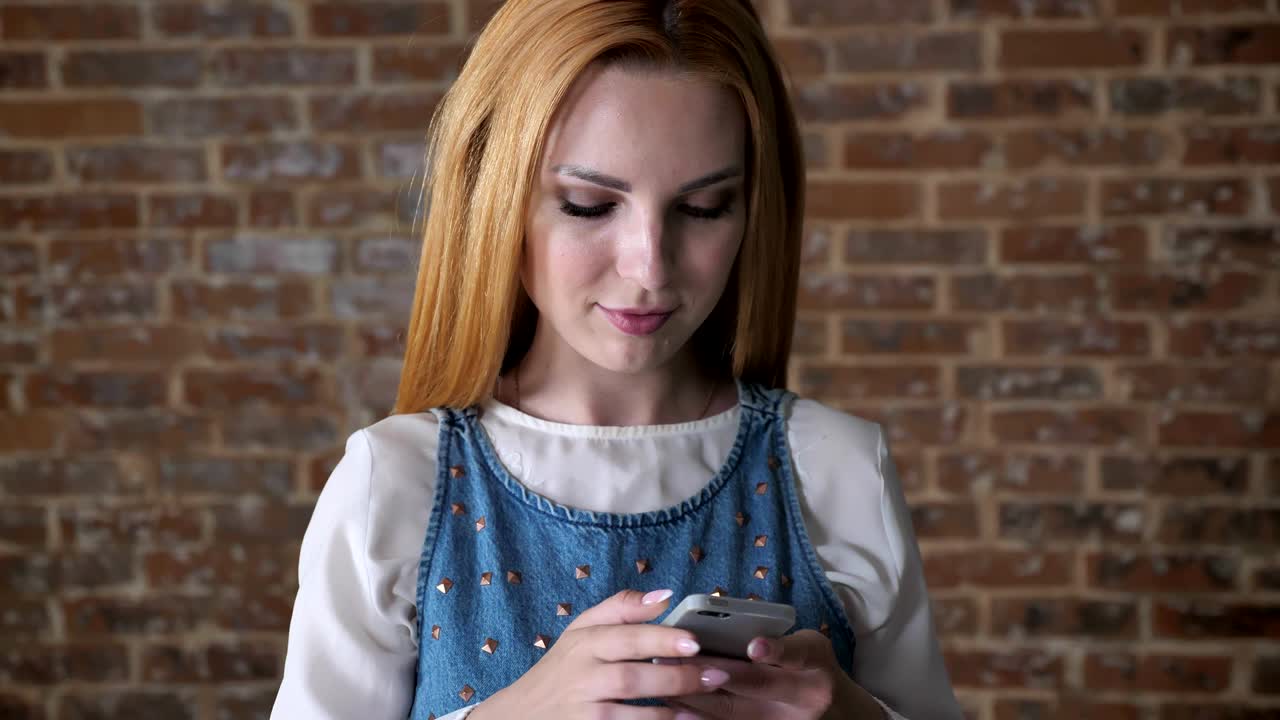 молодая соблазнительная блондинка печатает сообщение на смартфоне, кирпичный фон, концепция связи