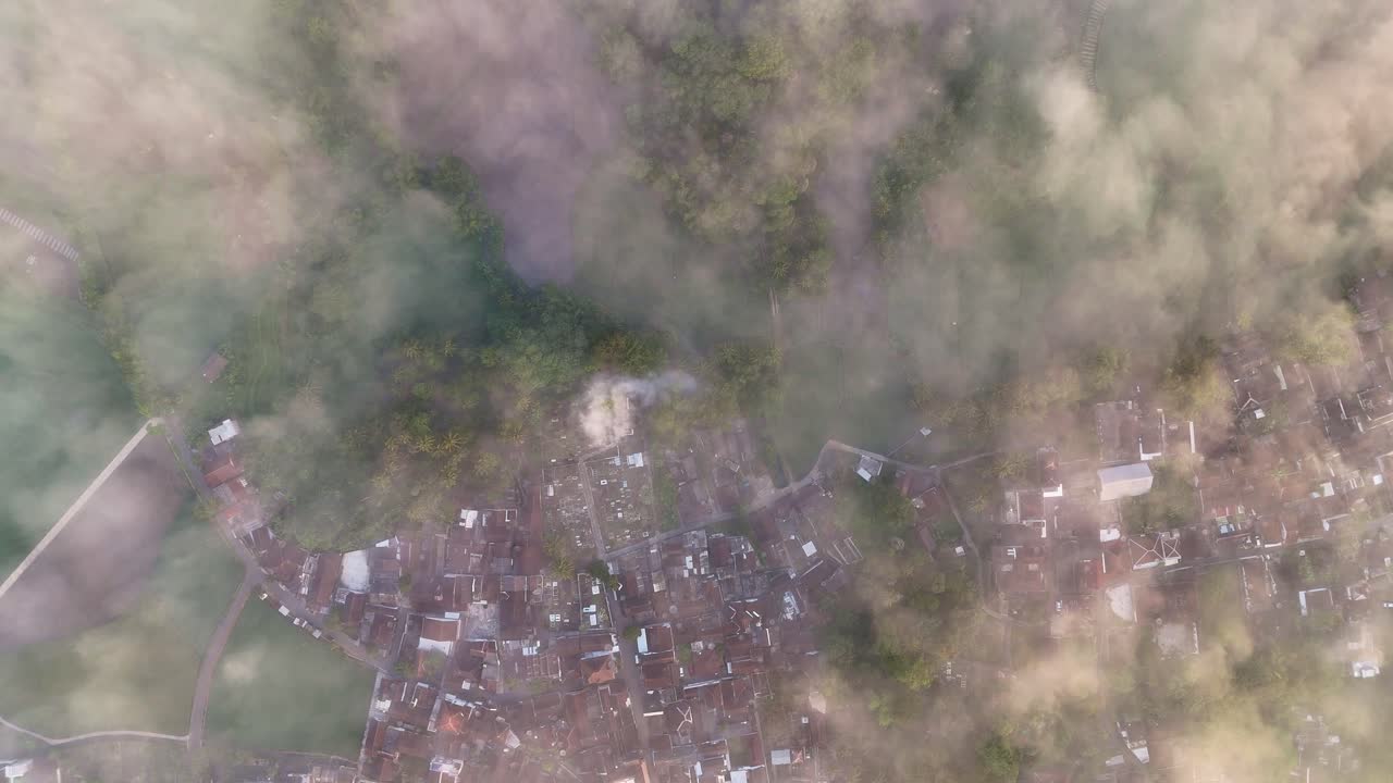 Aerial top-down view of a rural village partially covered in morning mist, surrounded by lush green forest and farmland, captured by drone in the morning