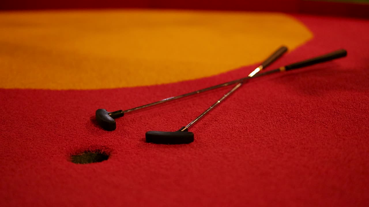 A vibrant orange golf ball rolls into a hole on a red and yellow miniature golf course under warm lighting