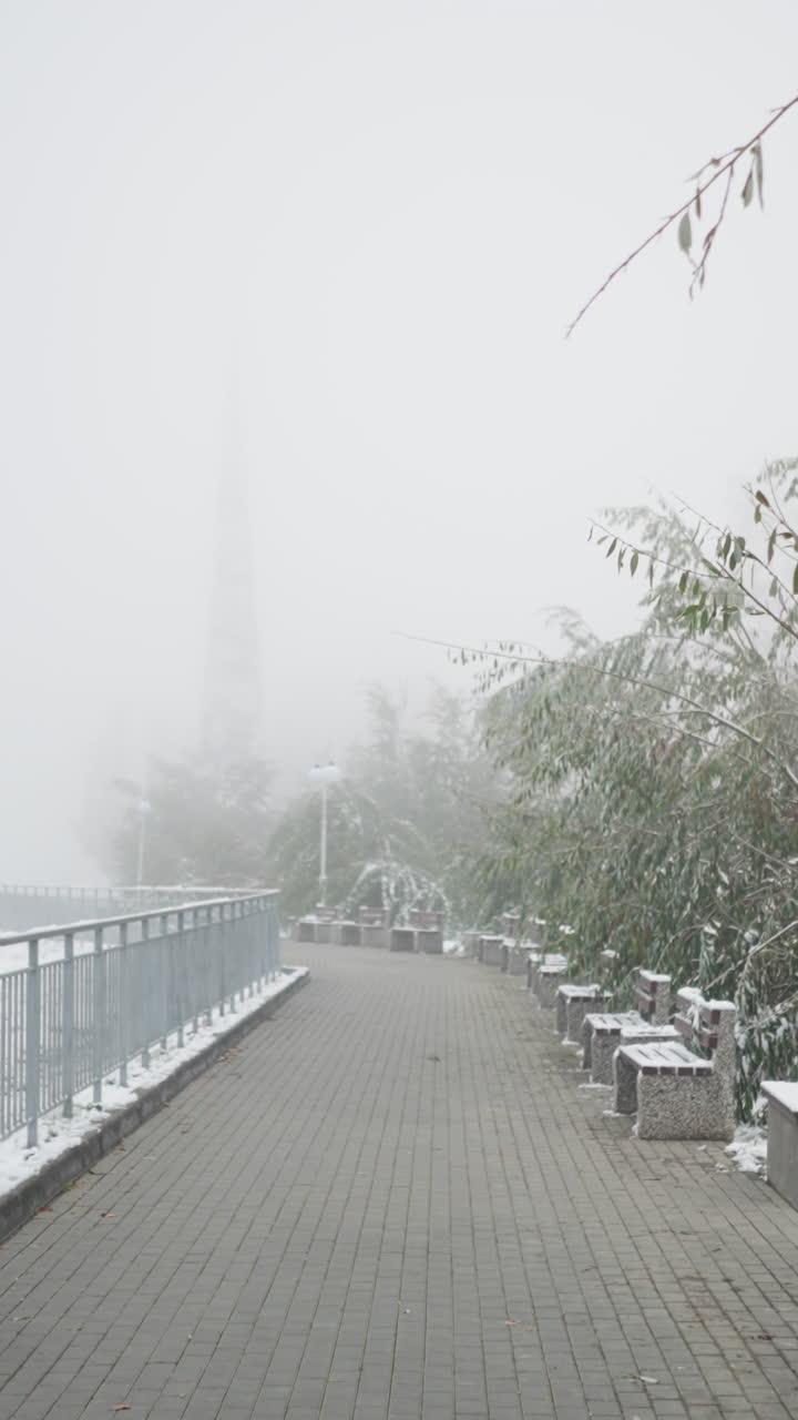 Early winter cityscape with first snowfall covering park walkway, benches, iron railings, and light poles, surrounded by frosted trees under foggy sky in quiet, muted, serene urban setting