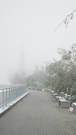 early winter cityscape with first snowfall covering park walkway, benches, iron railings, and light poles, surrounded by frosted trees under foggy sky in quiet, muted, serene urban setting