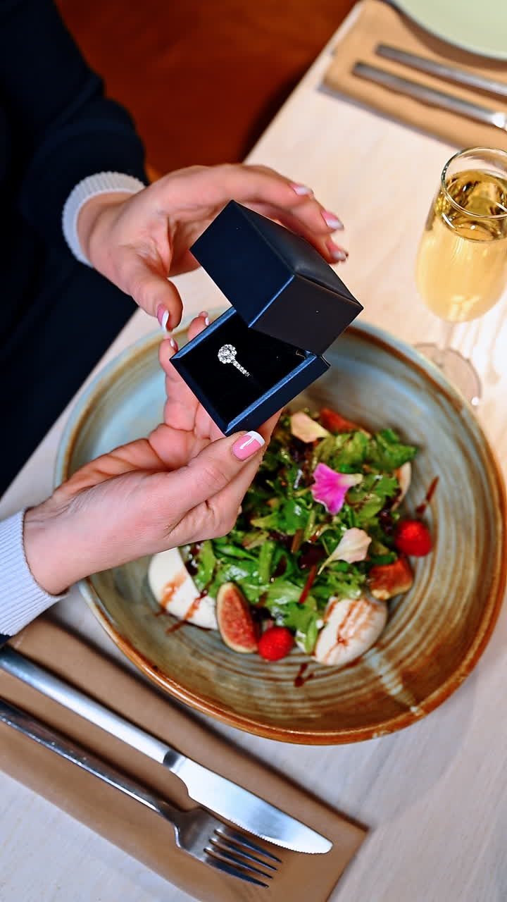 En person holder en ringæske over en salat under et særligt øjeblik på en restaurant. En person viser en ringæske, mens personen spiser en salat ved et restaurantbord. Dette øjeblik inkluderer spænding og overraskelse.
