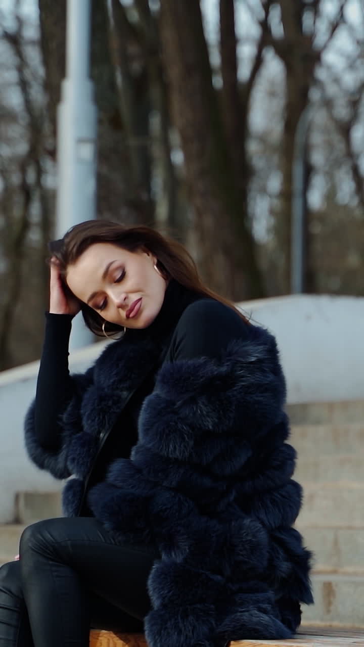 Luxury girl sitting on bench. Attractive young woman in fur coat enjoys fresh air in the park and smiles. Positive brunette outdoors. Vertical video