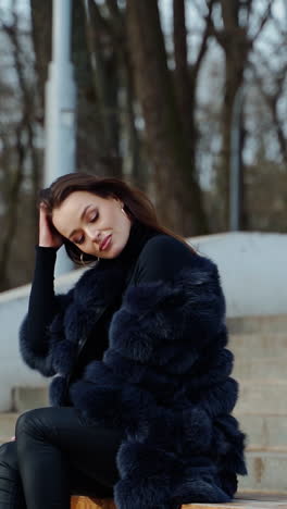 luxury girl sitting on bench. attractive young woman in fur coat enjoys fresh air in the park and smiles. positive brunette outdoors. vertical video