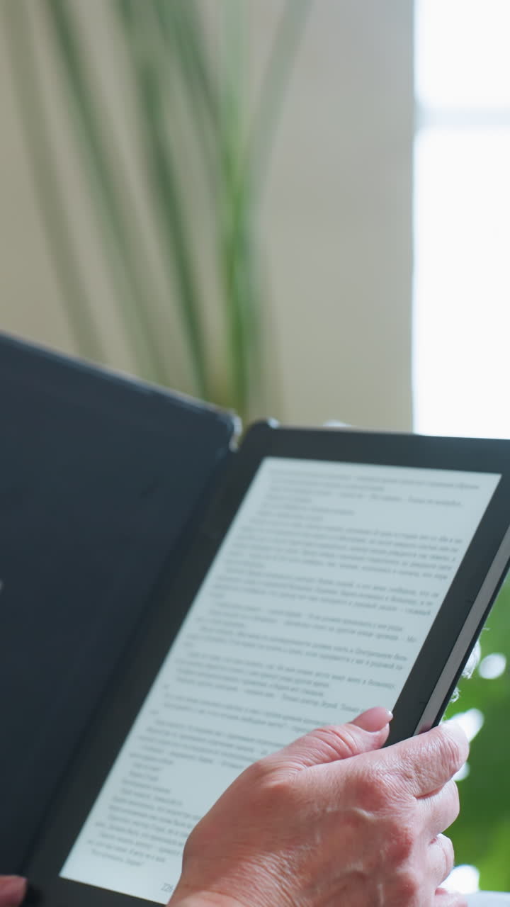 Mature woman using finger to scroll or turn page on digital e-reader while enjoying quiet reading moment indoors surrounded by green houseplants and soft natural light in peaceful cozy home setting