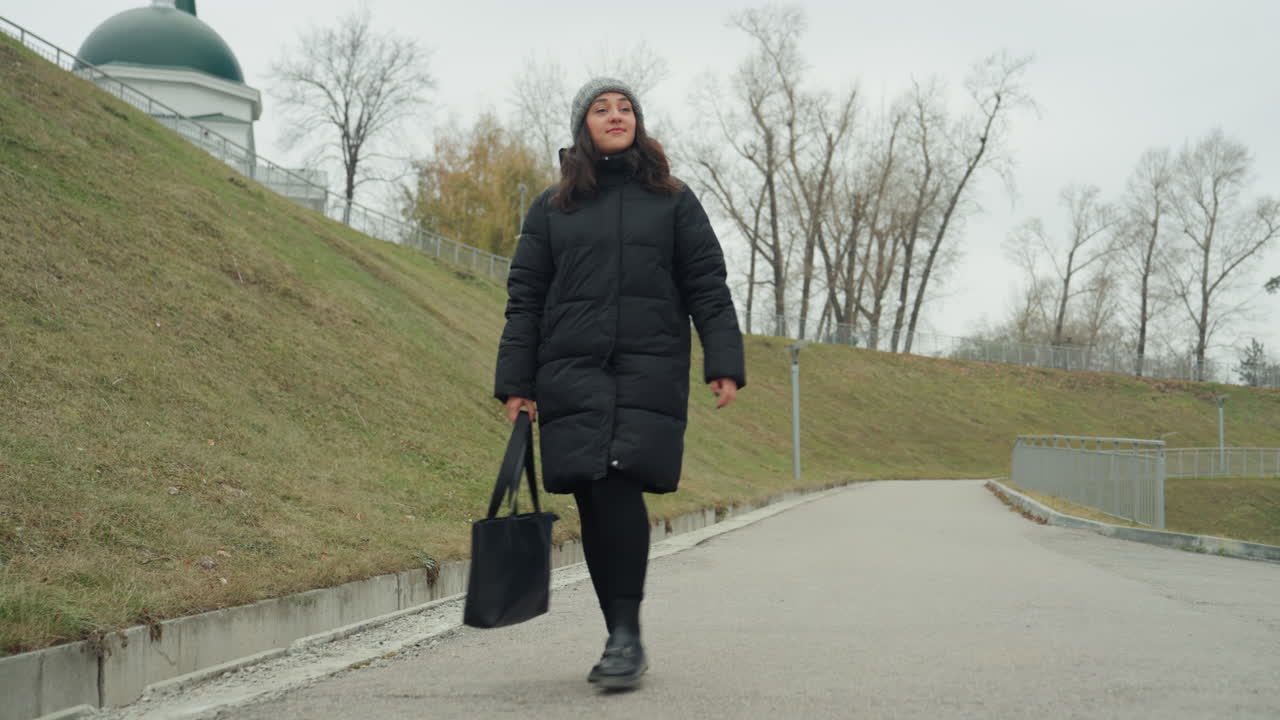 Signora calma in cappotto cammina su un sentiero deserto circondato da alberi sparsi. Una donna tranquilla, vestita con una giacca scura, si muove lentamente lungo un sentiero vuoto del parco con scarsa vegetazione e tonalità tenui.