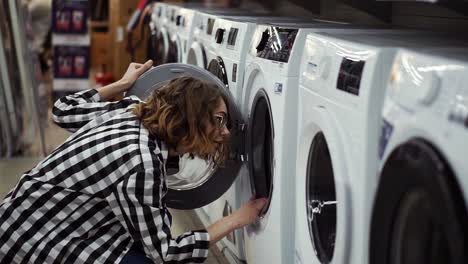 a young positive woman in a plaid shirt choosing washing machine in the shot of household appliances. opens the door and look