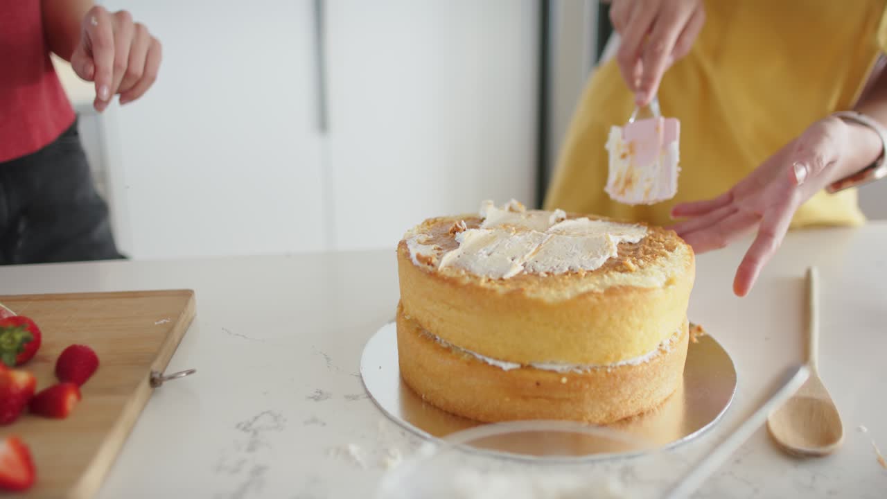2人の女性の友人が糖菓を味見し、キッチンのカウンターでヘラを使って2層のケーキを滑らかにしている様子