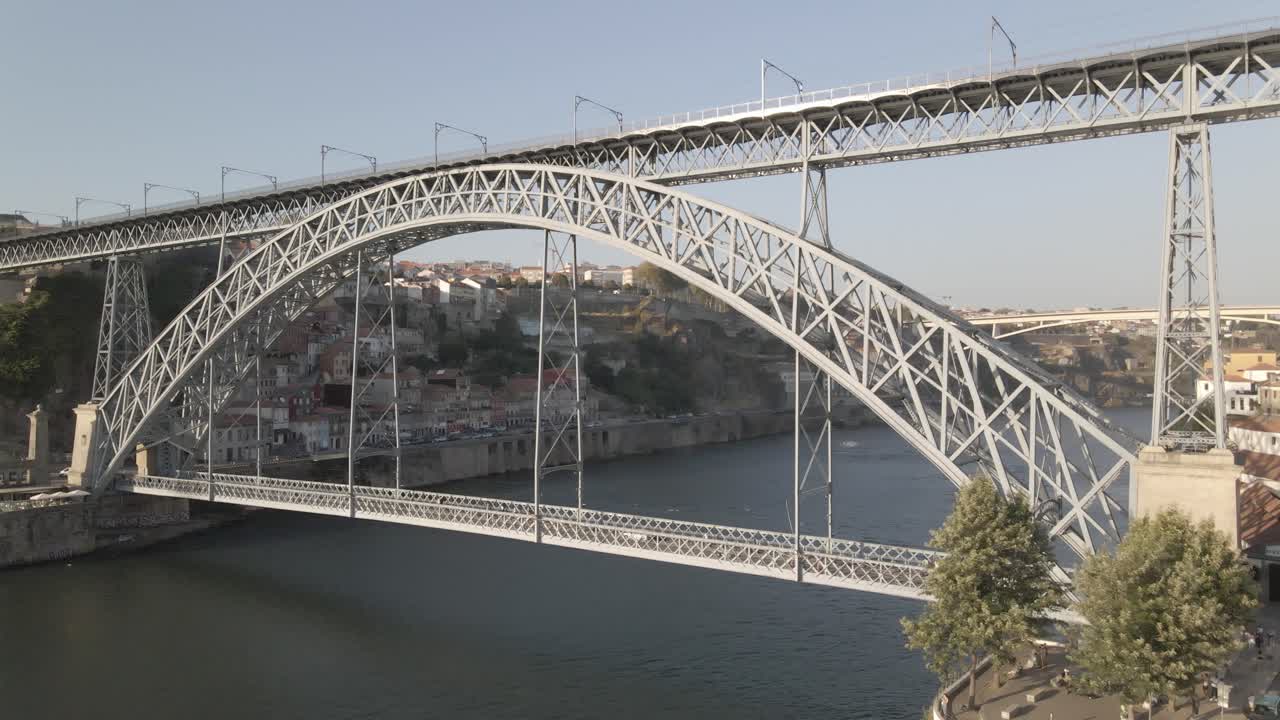 Aerial drone view of iconic Dom Luís I Bridge in Porto, Portugal, with double-deck iron arch spanning Douro River, connecting Porto and Vila Nova de Gaia under clear blue sky in daylight
