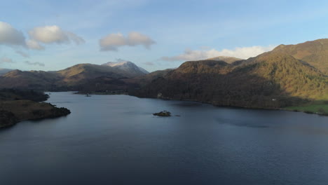 Hohe-Luftdrohne,-Die-über-Den-Ullswater-See-Mit-Schneebedecktem-Helvellyn-Berg-Im-Hintergrund-Geschossen-Wird,-Wird-Hügel-Mit-Bäumen-An-Einem-Sonnigen-Und-Bewölkten-Morgen-Im-Lake-District-Von-Cumbria,-Vereinigtes-Königreich