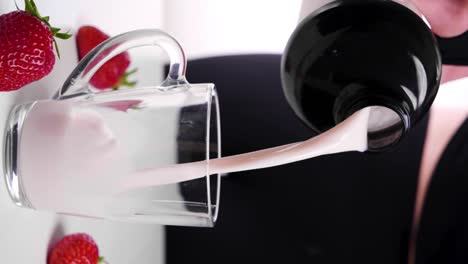 Tilt-slow-motion-shot-of-model-holding-shaker-with-strawberry-protein-shake,-filling-a-glass,-strawberries-on-table