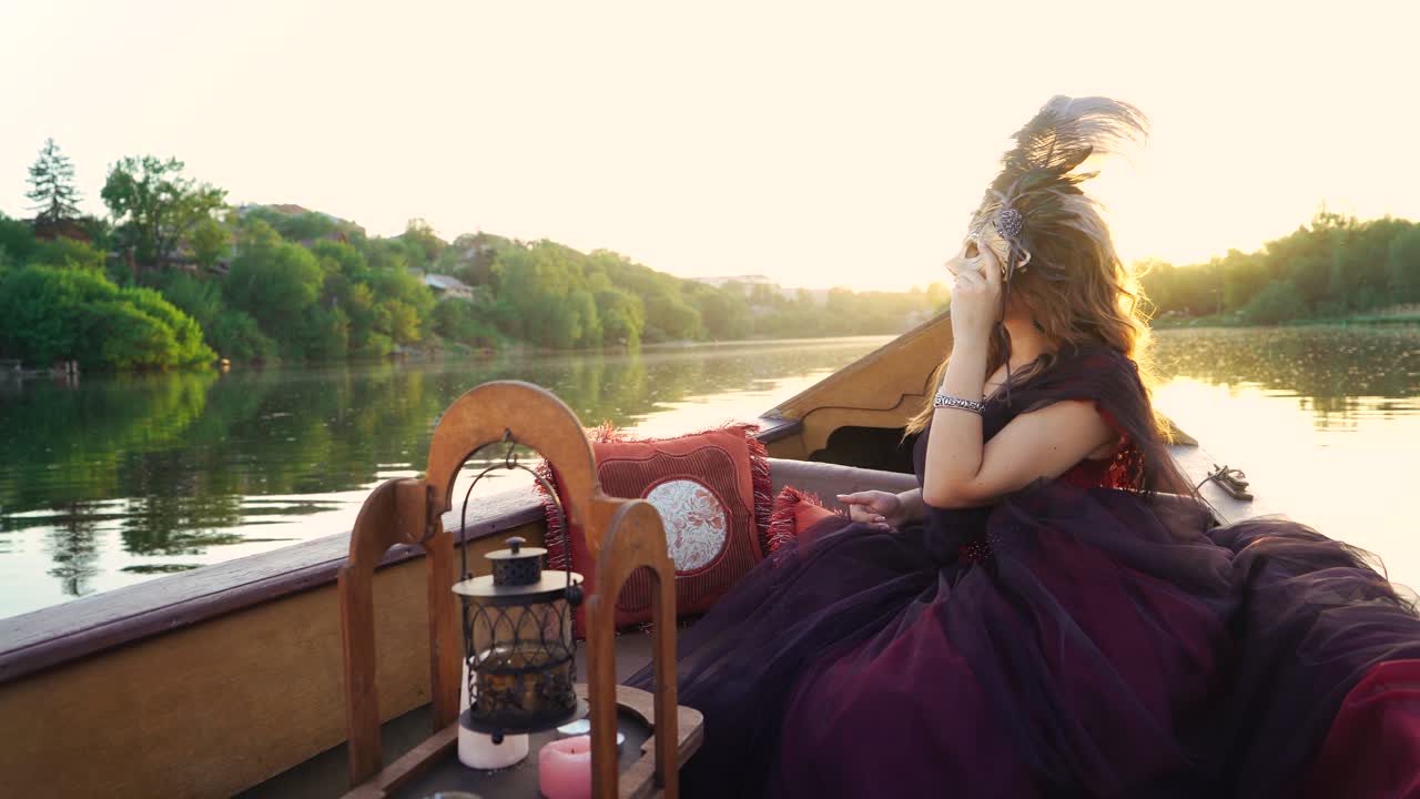Gorgeous female in masquerade costume sitting in gondola and looking at beautiful natural landscape. Woman holding carnival mask floating in a boat at sunset.