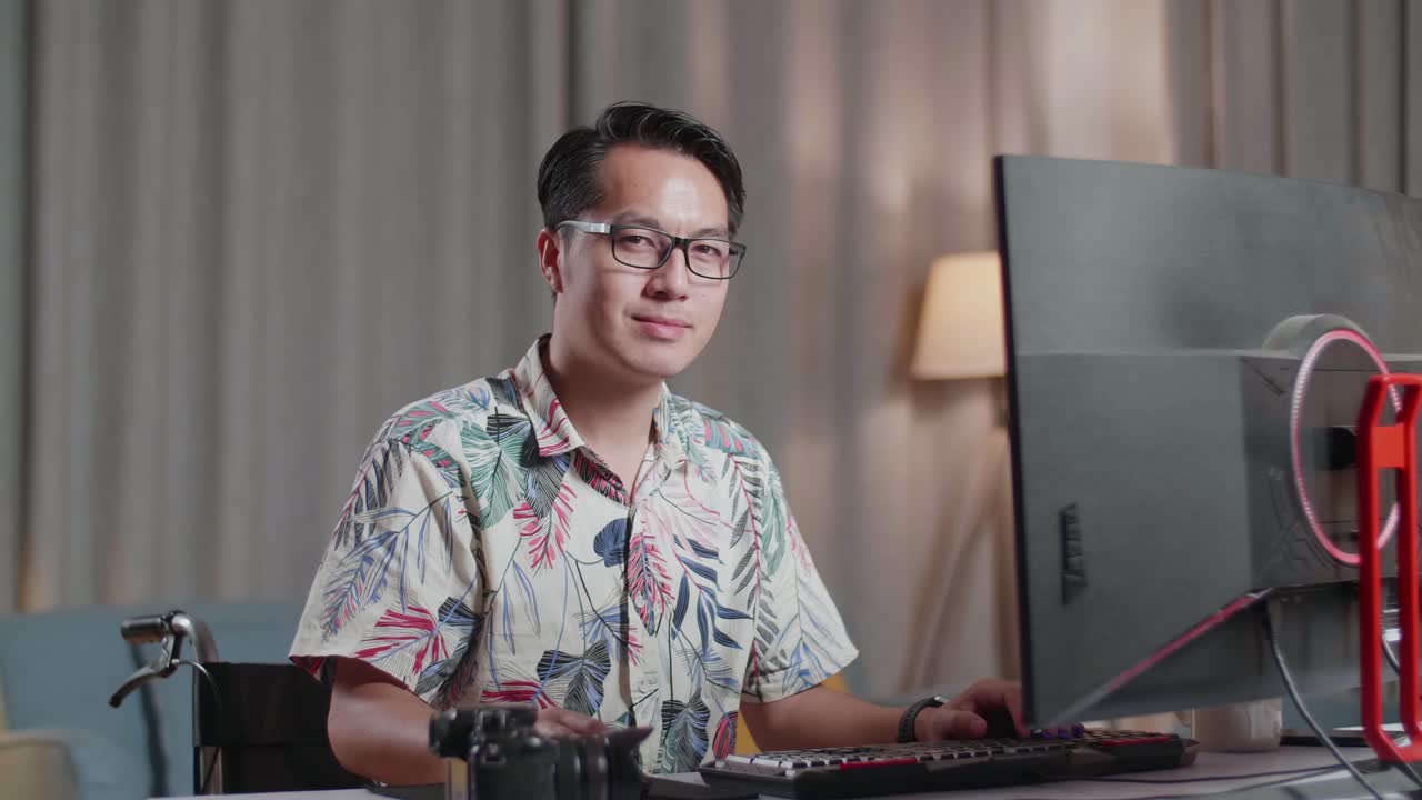 체어에 앉은 아시아인 남자가 미소 짓고 집에 있는 카메라 에 있는 데스크으로 비디오를 편집하는 동안 카메라로 팔을 고 있다.
