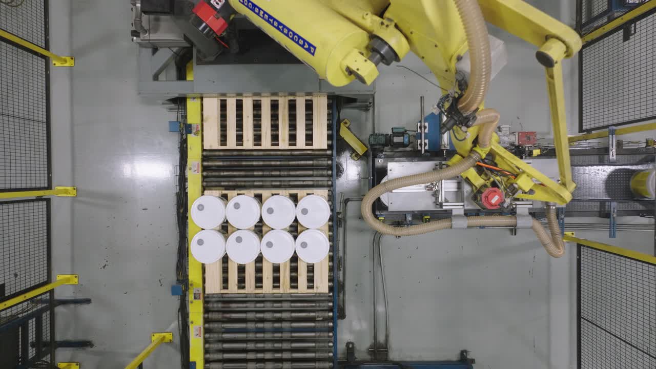 Top-down aerial shot of a yellow robotic arm performing palletizing by stacking white pails and buckets onto a wooden pallet on an automated production line