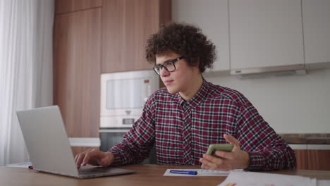 A-Curly-man-with-a-serious-look-works-at-a-laptop-sitting-in-a-modern-kitchen.-Young-man-freelancer-student-using-laptop-studying-online-working-from-home-in-internet