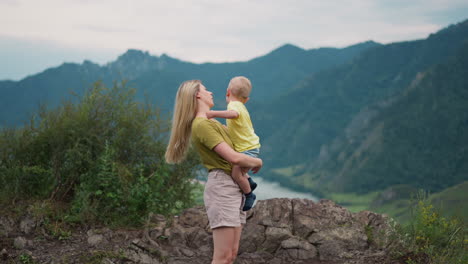 young mother adjusts loose hair holding toddler boy on hill