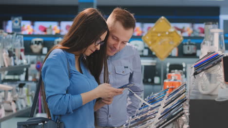 joven hermosa pareja casada un hombre con una camisa y una mujer con gafas sosteniendo un teléfono inteligente de pie alrededor de una vitrina con teléfonos inteligentes en una tienda de electrónica comprobando la pantalla táctil de la pantalla e inspeccionando la forma y el diseño del dispositivo