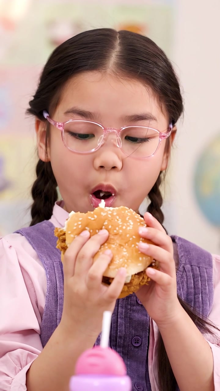 Young girl with glasses eats large chicken burger in bright classroom, static close-up shot