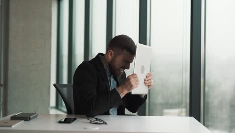 an angry man hits himself with a laptop while sitting behind a chair in a transparent office with panoramic windows