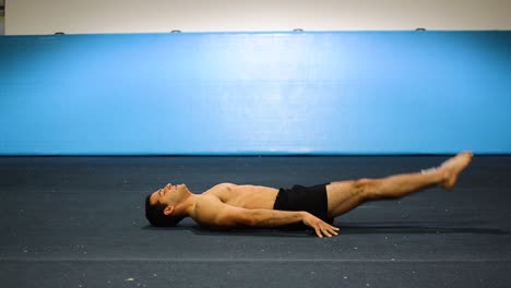 a guy doing an abs workout in a gymnastics gym doing lying down shortless leg and hand raises still shot