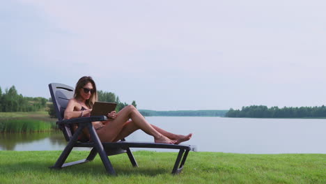 Woman-lying-on-a-sun-lounger-in-a-swimsuit-with-a-tablet-computer-finger-touches-the-screen-of-the-tablet-and-smiles-lying-on-the-lake