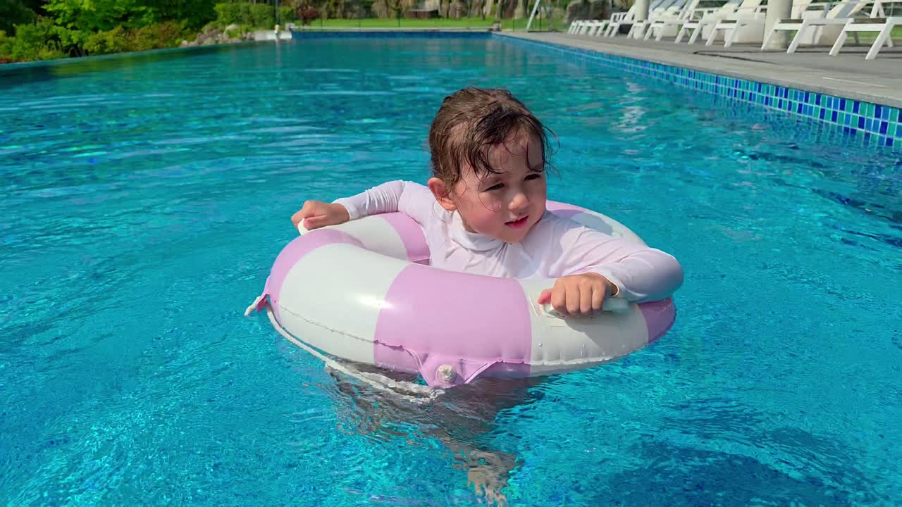 A joyful little girl in a rash guard laughs and has fun as she spins herself around in a pink inflatable ring in a bright blue swimming pool on a sunny summer day