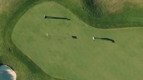 Vista-Aérea-De-Arriba-Hacia-Abajo-De-Dos-Golfistas-Masculinos-Y-Un-Palo-De-Bandera-Con-Largas-Sombras-Poniendo-Una-Pelota-De-Golf-En-El-Hoyo-En-Una-Tarde-Soleada-En-Un-Campo-De-Golf-De-Florida