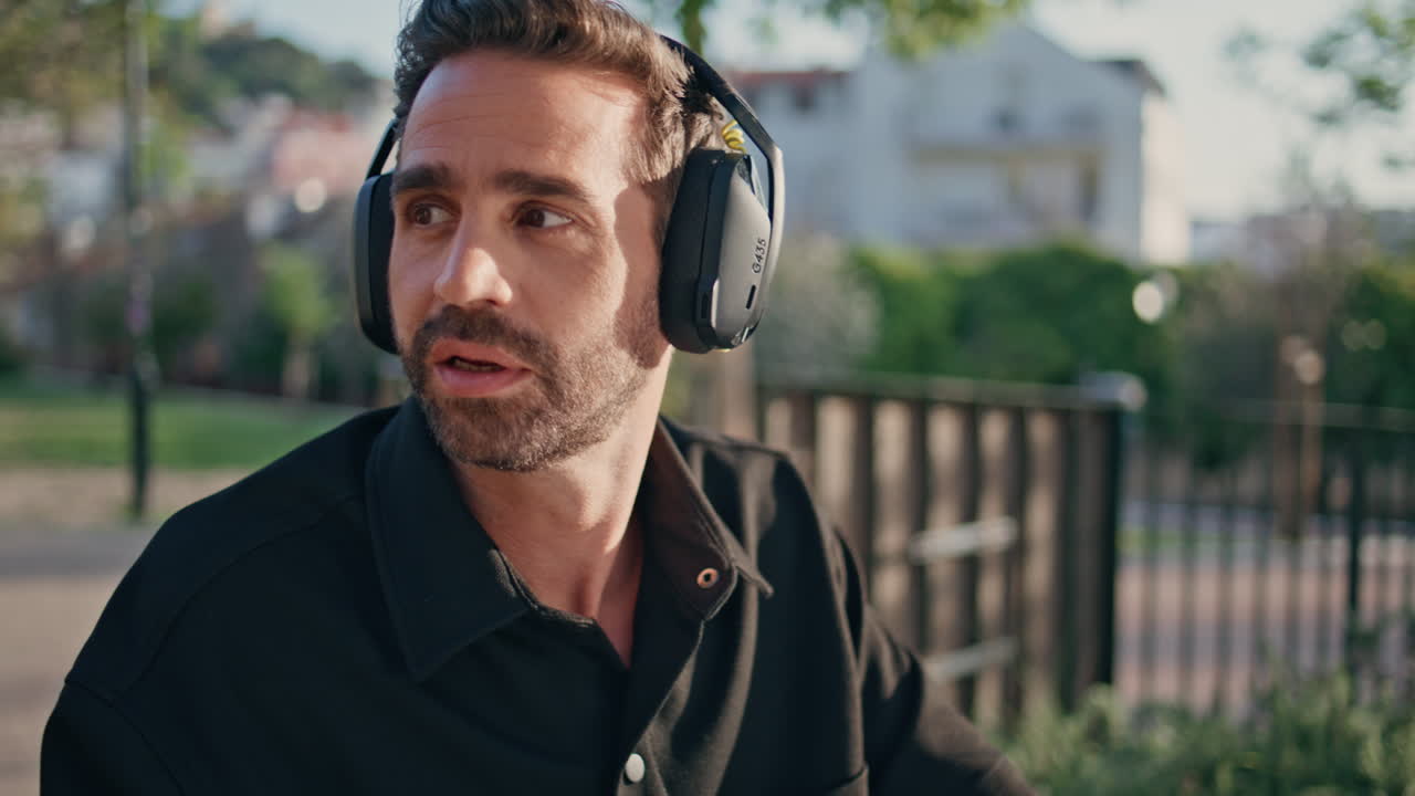 Headphones guy listening music sitting sunlight city park close up. Man headset