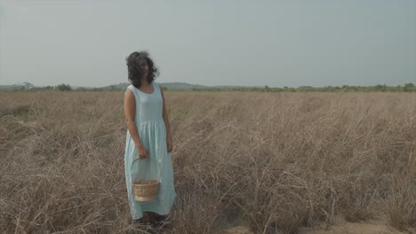 static slow motion shot of a young pretty indian woman in a light blue dress and a wood basket in her right hand standing on a dry farmer field stroking her hair as the wind blows past her