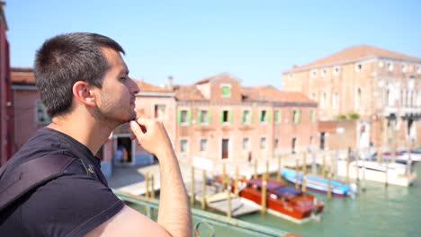 Young-tourist-man-looking-at-boats-over-the-canal-from-a-bridge-in-Murano,-Venice