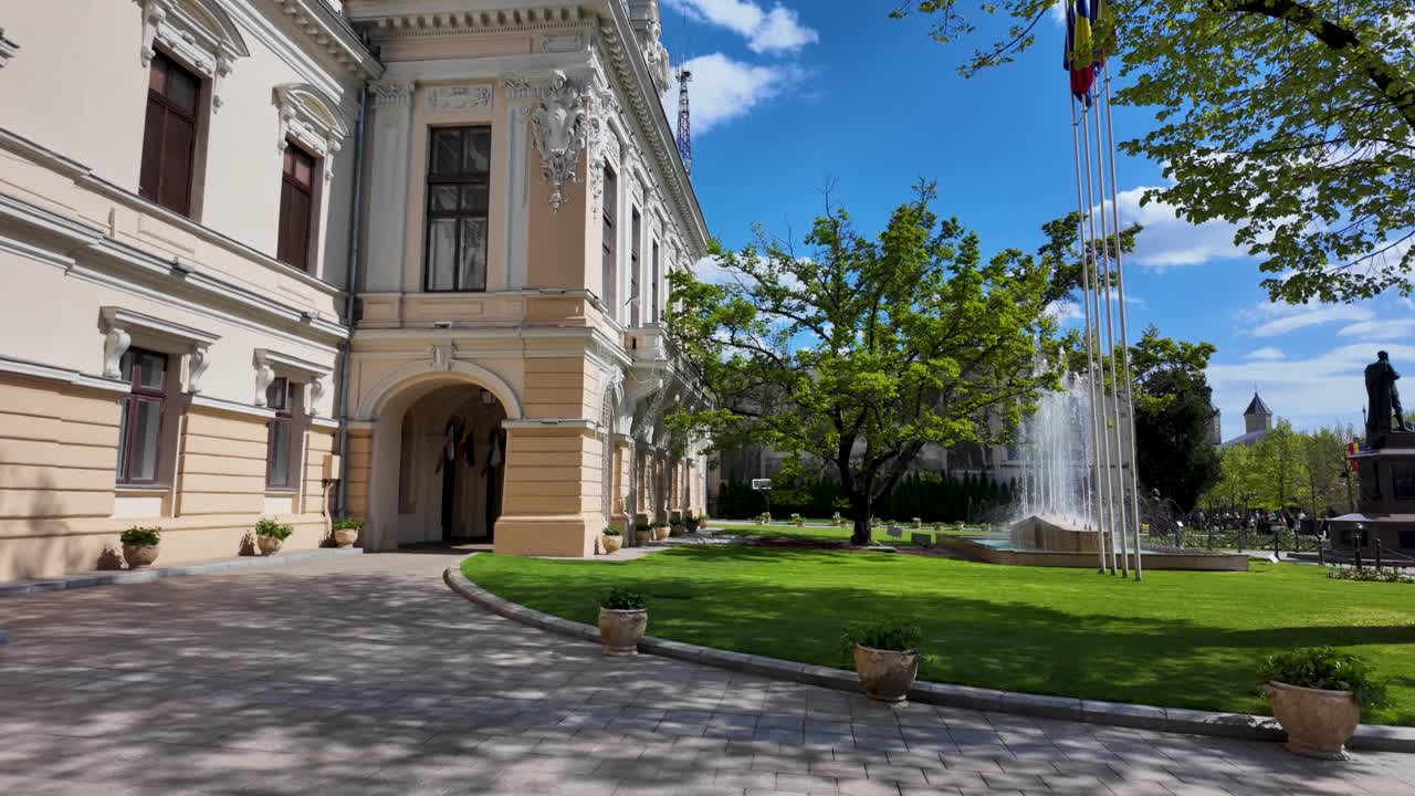 Vista en cámara lenta del patio del Ayuntamiento de Iasi en Stefan cel Mare con un árbol, una fuente y banderas en un día soleado
