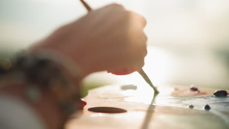 a close-up of a woman's hand, adorned with beads, as she mixes colors on a palette with a brush under soft sunlight. the scene highlights the delicate and focused process of blending paints