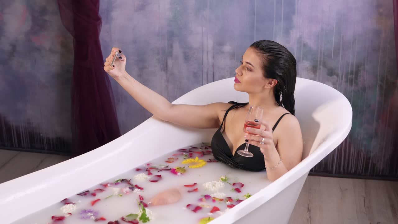아름다운 젊은 여성이 우유 욕조에서 휴식을 취하고 잔에서 스파클링 와인을 마시며 셀카를 찍고 있습니다. 4k로 촬영되었습니다.