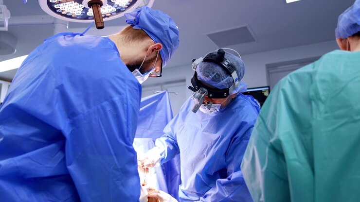 Kyiv, Ukraine, 5 May 2026: Surgical team with headlamp loupe operating under LED surgical lamp. Surgeons in blue gowns focus on operative field, round LED light and monitor visible behind.