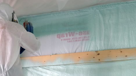 worker in respirator and tyvek suit sprays first layer of closed cell foam insulation near the ceiling of a newly constructed exterior wall