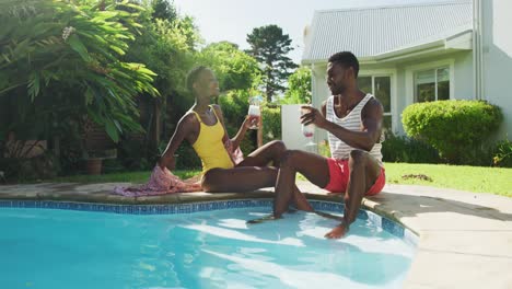 happy african american couple sitting by swimming pool making a toast with drinks in sunny garden