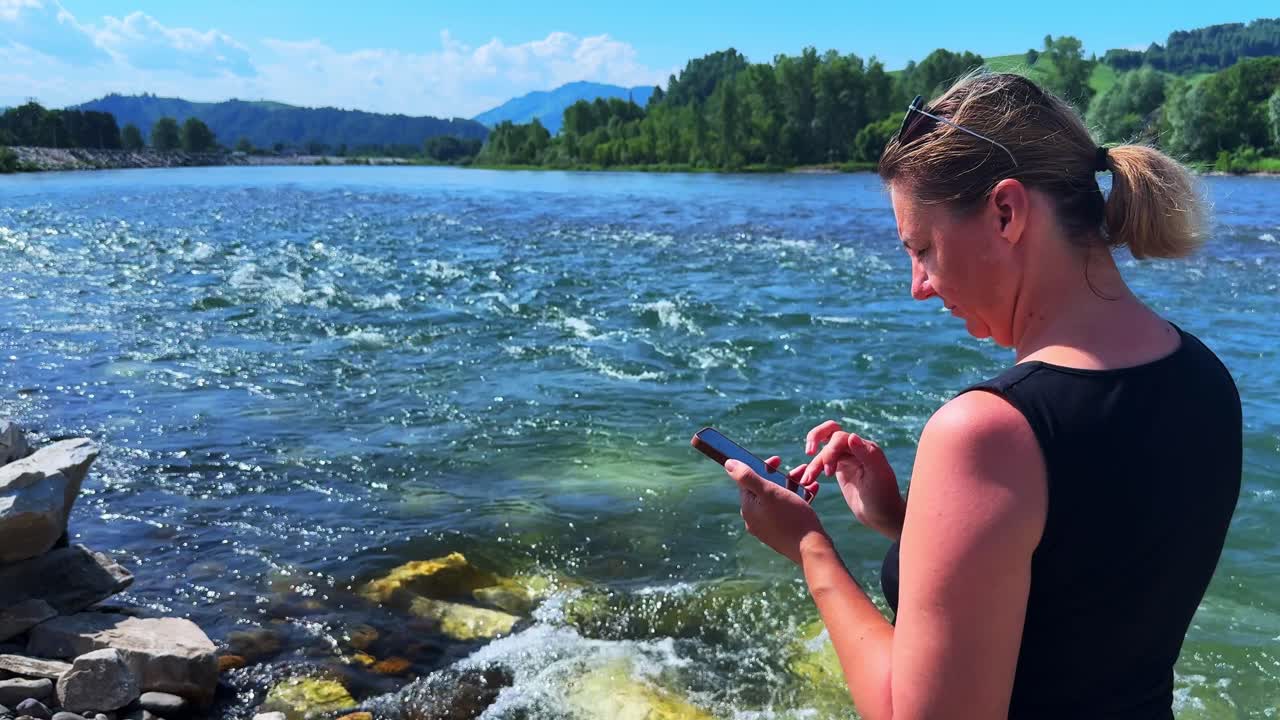 A Person Engaging with Their Smartphone While Enjoying a Scenic River View Surrounded by Lush Greenery and Mountainous Backgrounds Under a Clear Blue Sky