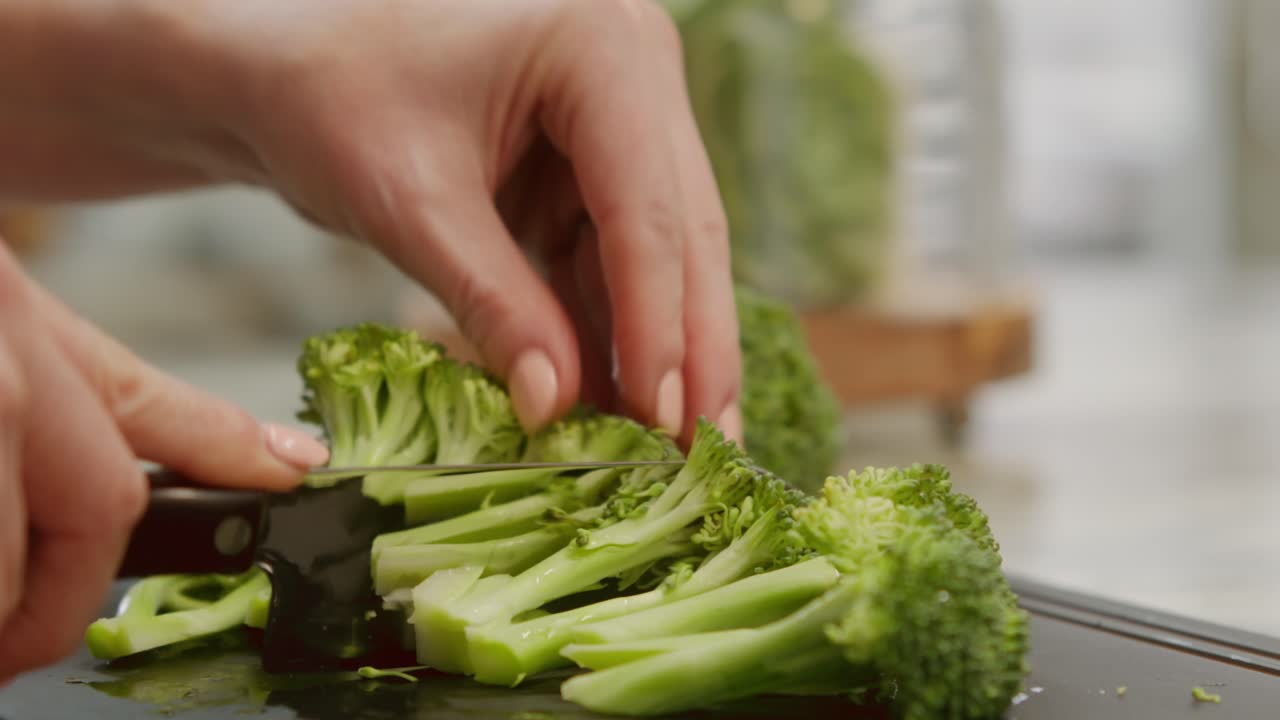 Caucasian hands slice stems of broccoli vertically on a cutting board