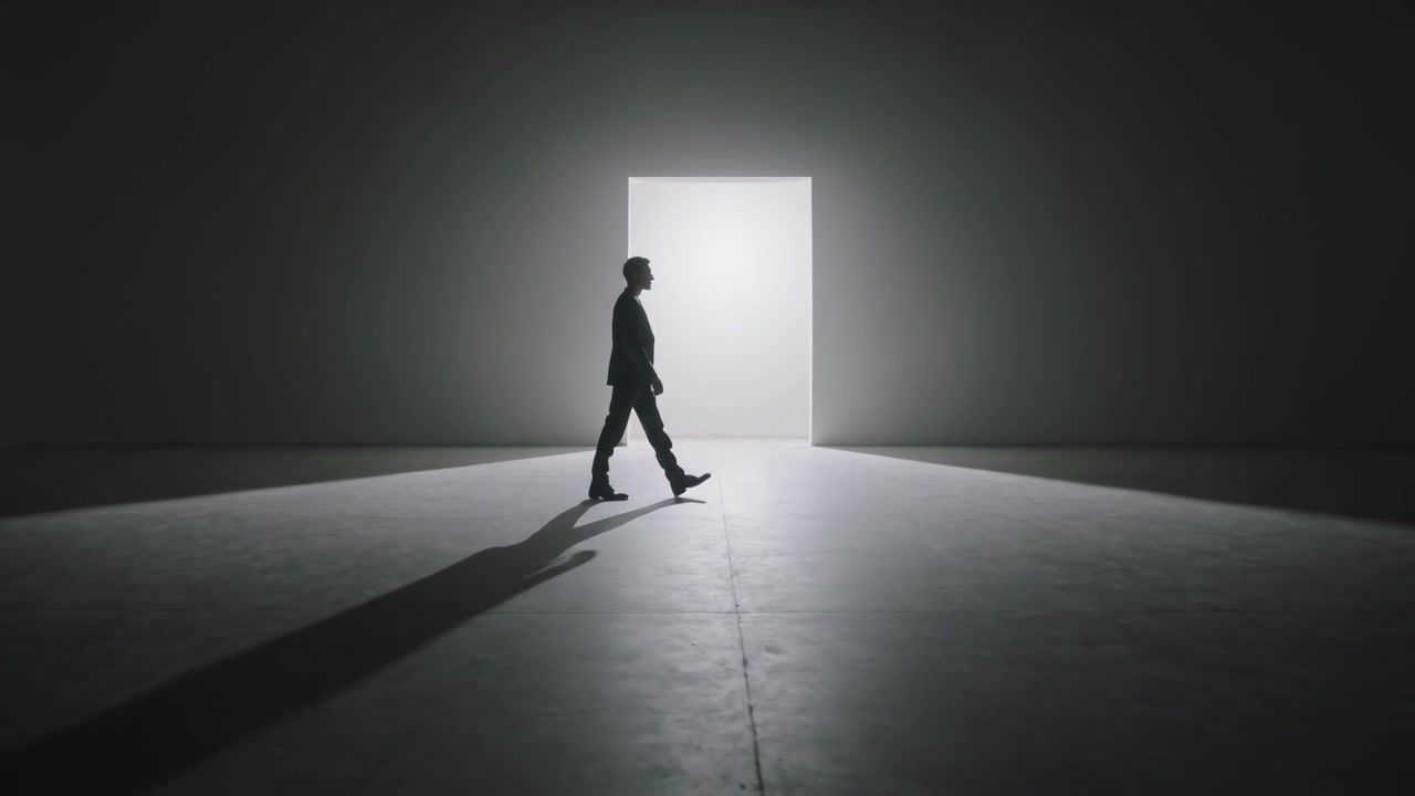 Entrando da sinistra, un uomo in abito cammina verso un portale luminoso in un interno fioco, con l'ombra che lo segue.